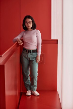 Minimal full length portrait of Asian young woman standing in statement red space and looking at camera