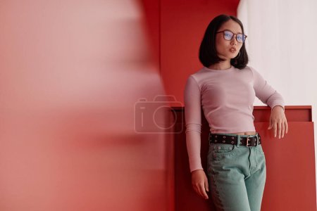 Minimal fashion portrait of confident Asian woman wearing eyeglasses and looking at camera standing in statement red space, copy space