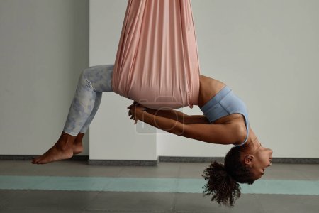 Retrato de vista lateral de una joven negra disfrutando de yoga aéreo y relajante estiramiento en hamaca rosa