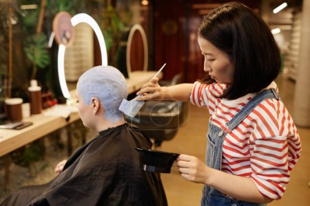 Retrato de vista lateral de mujer asiática joven como estilista que aplica lejía al pelo corto de corte de zumbido en el salón de belleza