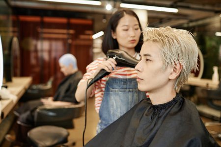 Seitenansicht Porträt eines jungen asiatischen Mannes im Schönheitssalon mit Friseur, der gebleichte Haare trocknet, Kopierraum