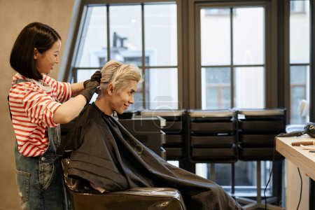 Side view portrait of smiling young Asian man in beauty salon with hairstylist applying bleach to hair, copy space