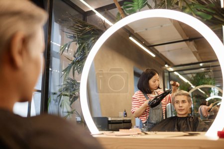Mirror reflection shot of hairstylist drying and styling hair of young Asian man in beauty salon, copy space