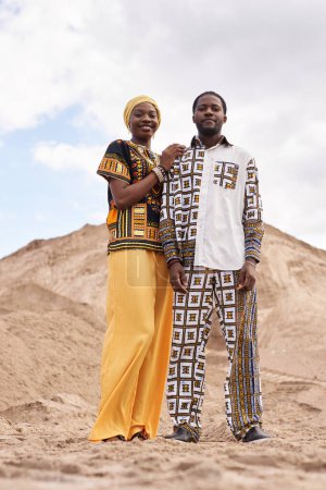 Vertical full length portrait of young Black couple wearing traditional clothing posing in desert landscape and looking at camera