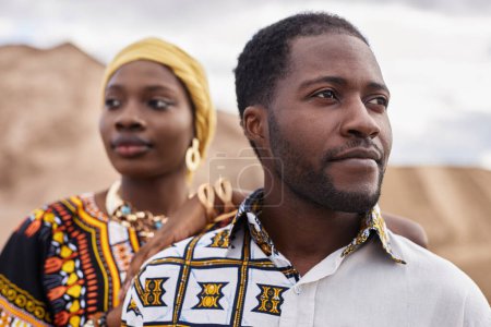 Fashion portrait of African American couple wearing traditional clothing and posing in desert, focus on man looking away in foreground