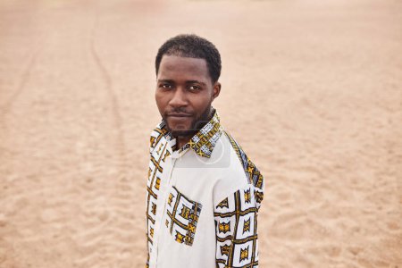 High angle portrait of African American man wearing traditional patterns looking at camera in desert, copy space