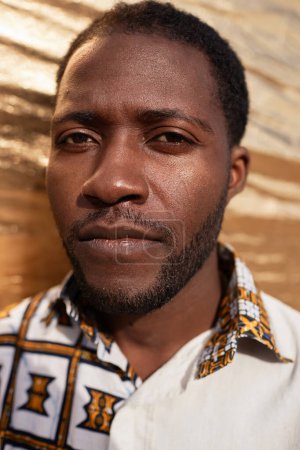Closeup portrait of Black man wearing traditional pattern in gold light and looking at camera with intense face expression