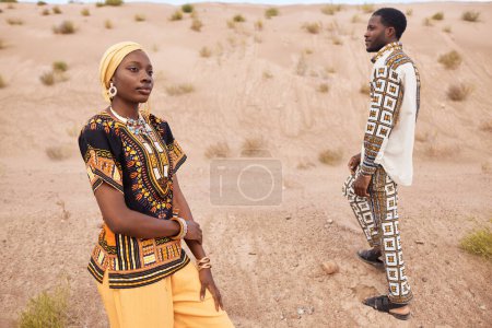 Fashion portrait of African American couple wearing traditional clothing posing in desert setting, focus on young woman wearing chunky jewelry in foreground