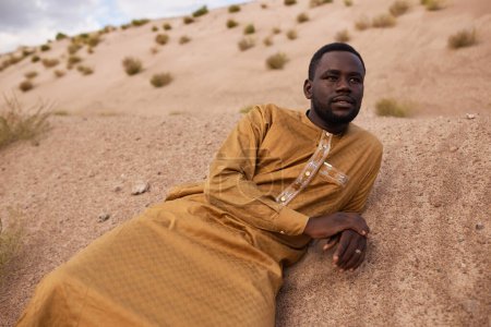 Porträt eines afrikanisch-amerikanischen Mannes in einem langen Kaftan-Kleid, der auf Sanddünen in der Wüste liegt und wehmütig wegblickt