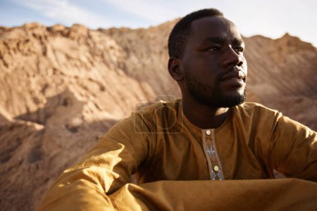Retrato de cerca del hombre negro sentado en la duna de arena en el desierto y mirando hacia otro lado bajo la dura luz del sol, espacio para copiar