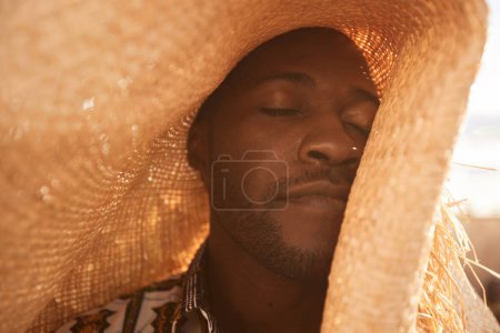 Retrato de cerca del joven negro con sombrero de paja y disfrutando de la luz del sol felizmente con los ojos cerrados, espacio para copiar