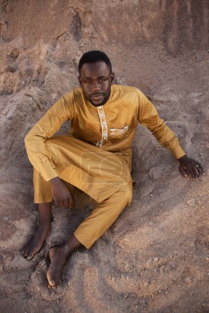 High angle portrait of Black young man wearing traditional kaftan sitting on sand in shadow and looking up at camera