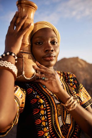 Vertical portrait of beautiful Black woman wearing traditional dress and carrying drum looking at camera outdoors in sunlight