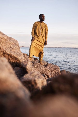 Retrato vertical del hombre negro tradicional caminando a lo largo de la costa rocosa del mar en la luz del atardecer