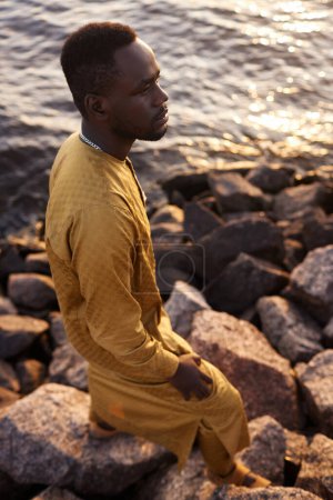 Vertical portrait of traditional Black man sitting on rocky sea shore in sunset light