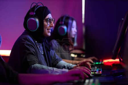 Photo for Side view portrait of Muslim young woman playing video games in cybersports club and smiling happily, copy space - Royalty Free Image
