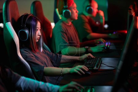 Photo for Side view portrait of young Asian woman playing video games in cybersports club and wearing pro headphones, copy space - Royalty Free Image