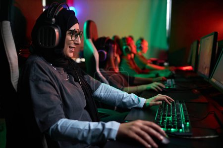 Photo for Side view portrait of smiling Muslim woman playing video games in cybersports club with neon lighting, copy space - Royalty Free Image