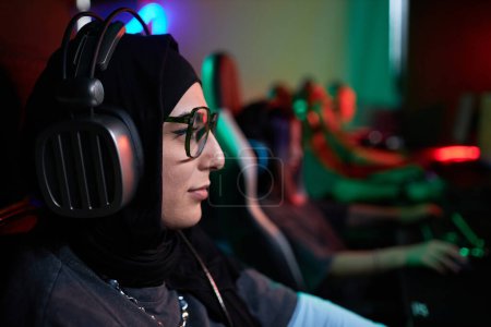 Photo for Side view closeup of young Muslim woman playing video games in cybersports club and wearing pro headphones, copy space - Royalty Free Image