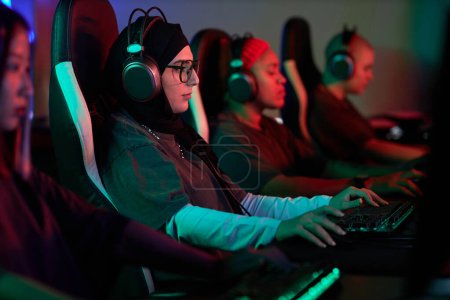Photo for Side view portrait of young Muslim woman playing video games with professional team in cybersports club, copy space - Royalty Free Image