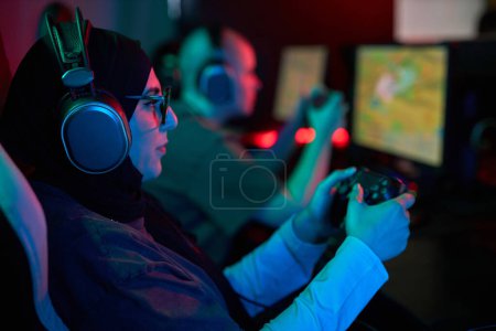 Photo for Side view portrait of Muslim young woman playing videogames in cybersports club and holding gaming controller, copy space - Royalty Free Image