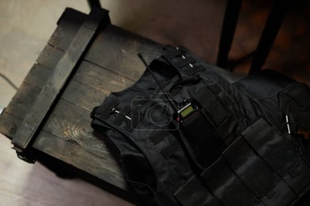 Above angle of black protective bulletproof body armor with walkie-talkie in pocket lying on wooden chest in military office
