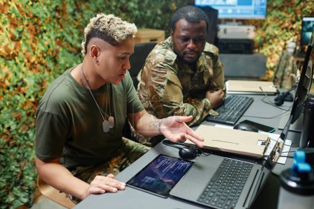 Young multiethnic female military officer pointing at laptop screen and explaining data to African American male colleague by workplace