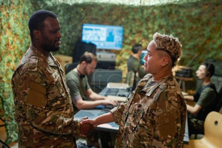 Two young intercultural officers in camouflage uniform shaking hands and looking at one another while standing in command and control center