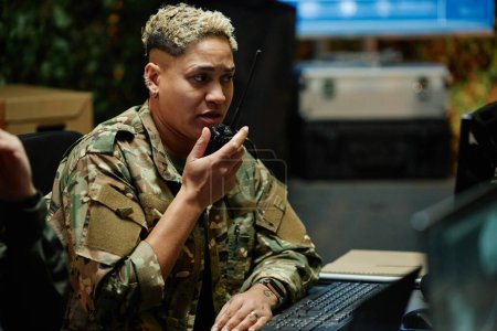 Young multiethnic female officer of surveillance center using walkie-talkie while reporting something and looking at computer screen