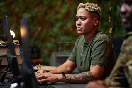 Young serious multiethnic female military officer keeping fingertips on computer keyboard and looking at screen by workplace