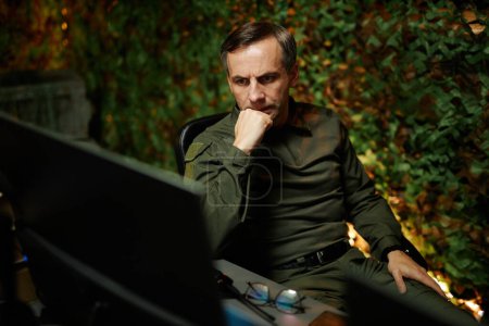 Serious or pensive mature male security or officer looking at computer screen while sitting by workplace in surveillance room