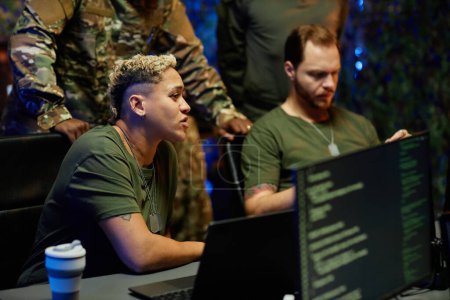 Young multiethnic female military officer sitting by workplace among male colleagues and looking at computer screen with surveillance camera