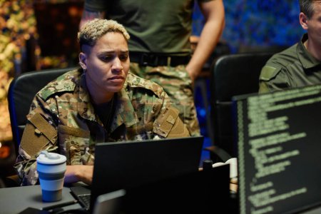 Young multiethnic female military officer in camouflage uniform looking through data on laptop screen while working among male colleagues