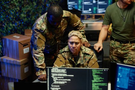 Serious female officer in camouflage uniform looking at computer screen while sitting between two young intercultural male colleagues