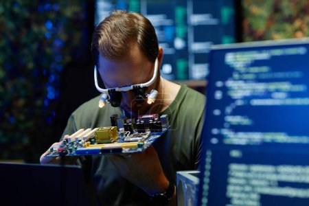 Young military officer or repairman wearing eyewear with small lamps checking details of processor among computer screens with data