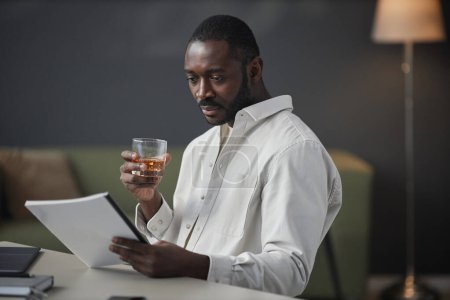 Photo for Side view portrait of successful African American entrepreneur reading documents at workplace in office and holding glass of whiskey, copy space - Royalty Free Image