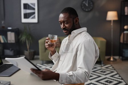Photo for Side view portrait of successful African American entrepreneur holding glass of whiskey while reading documents at workplace in office, copy space - Royalty Free Image