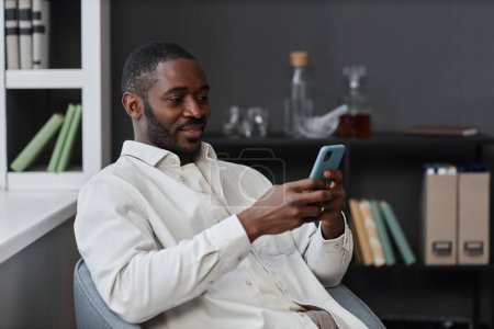 Photo for Portrait of adult African American man using smartphone relaxing in chair and scrolling social media, copy space - Royalty Free Image