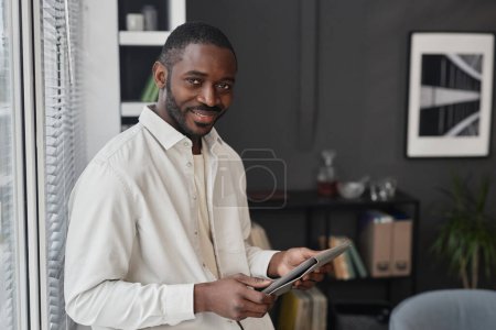 Photo for Waist up portrait of successful African American man smiling at camera standing by window with tablet, copy space - Royalty Free Image