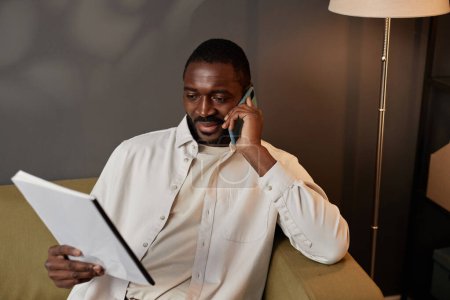 Photo for Portrait of successful Black man reading document and speaking on phone in office against gray wall, copy space - Royalty Free Image