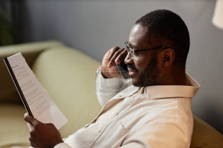 Photo for Side view portrait of successful Black man speaking on phone and reading document relaxing in office, copy space - Royalty Free Image