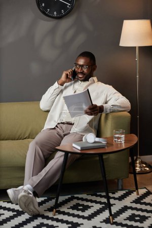 Photo for Vertical full length portrait of successful African American man speaking on phone relaxing on couch in office - Royalty Free Image