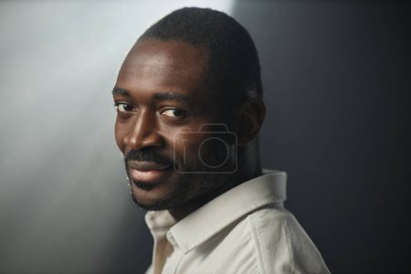Photo for Dramatic closeup portrait of African American adult man smiling at camera over shoulder on black background, copy space - Royalty Free Image