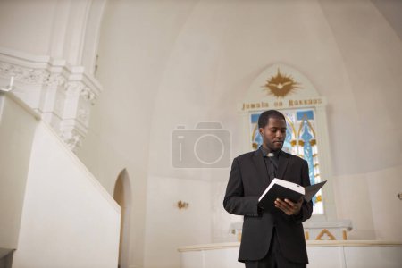 Foto de Retrato de gran angular del hombre afroamericano como sacerdote leyendo la Biblia en el altar de la iglesia, espacio para copiar - Imagen libre de derechos