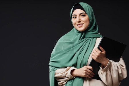 Téléchargez les photos : Portrait taille haute de jeune femme du Moyen-Orient tenant un ordinateur portable et souriant à la caméra debout sur fond noir en studio, espace de copie - en image libre de droit