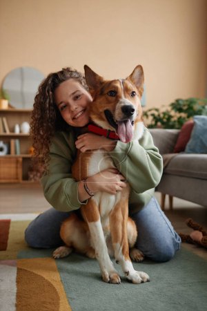 Full length portrait of happy young woman embracing big dog sitting on floor in cozy home