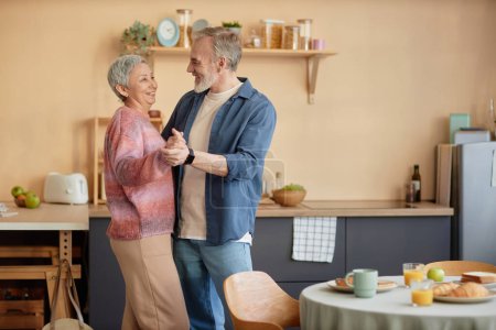 Side view portrait of happy senior couple slow dancing together in cozy kitchen and enjoying retirement copy space