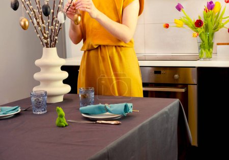 Foto de Mujer en vestido amarillo cuelga huevos de Pascua en ramas de sauce. Árbol de Pascua decorado o decoración casera DIY. Vacaciones religiosas de primavera. - Imagen libre de derechos