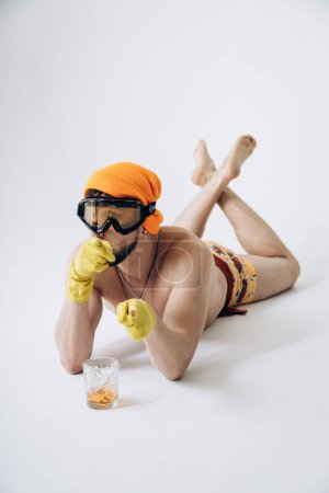 Photo for Guy in shorts in hat and ski goggles, on his hands yellow rubber gloves lies on floor sunbathing - Royalty Free Image