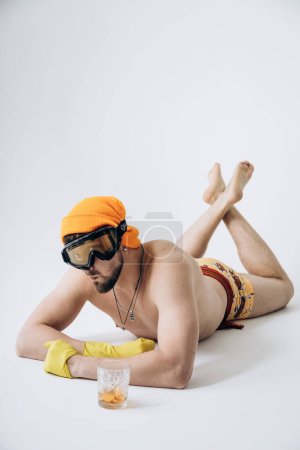 Photo for Guy in shorts in hat and ski goggles, on his hands yellow rubber gloves lies on floor sunbathing - Royalty Free Image